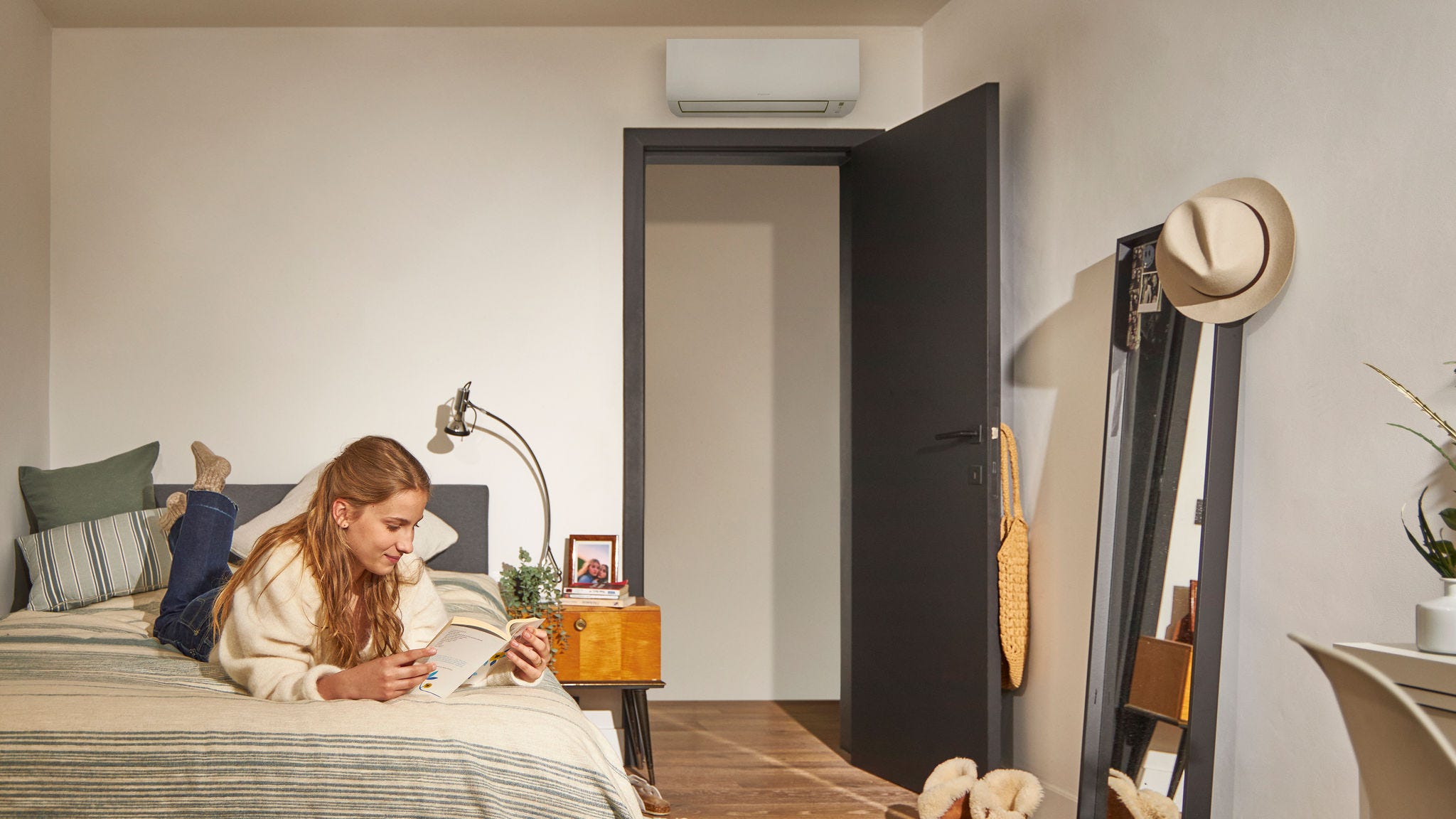 Een vrouw leest op een bed in een gezellige slaapkamer met een warmtepomp airconditioner boven de deur, die zorgt voor energiezuinige klimaatbeheersing
