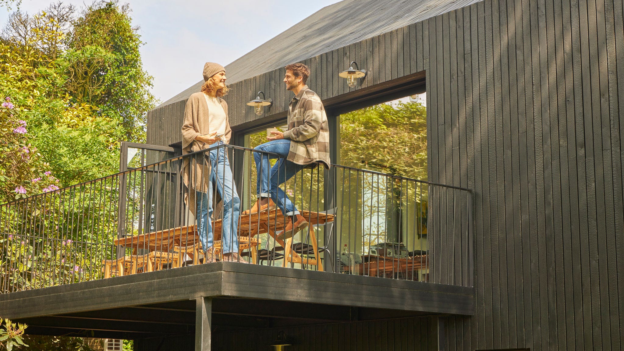Een koppel is aan het kletsen op het terras van een moderne, houten bungalow, terwijl het omringd is door natuur en geniet van frisse lucht en energiezuinige leefomstandigheden