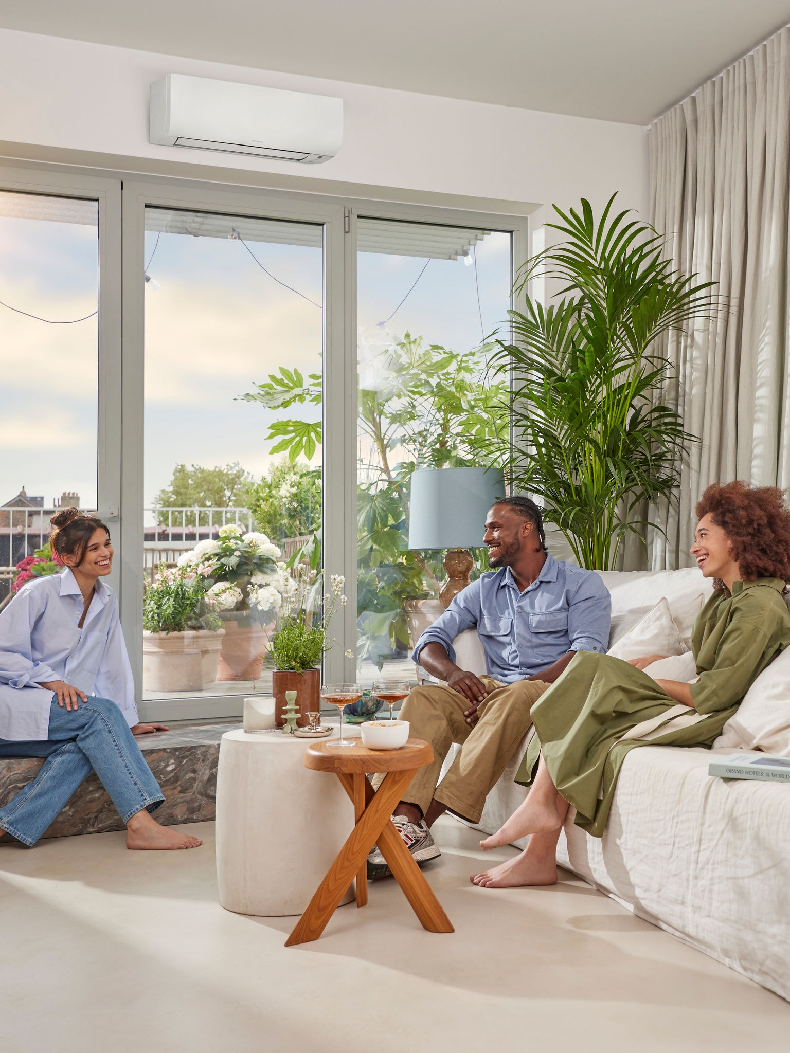 Een man en twee vrouwen zitten gezellig te praten in een lichte ruimte met witte muren en een onopvallend wit wandmodel warmtepomp boven een groot raam op een zonnige dag