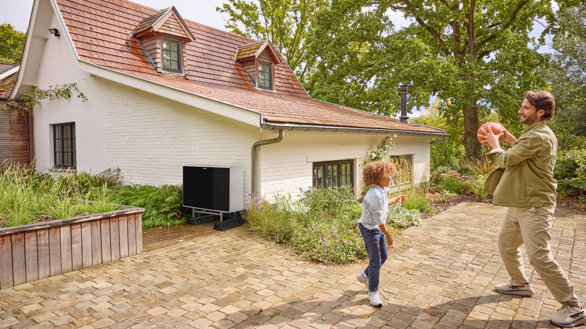 Vader die buiten in de tuin met zijn kind een bal overgooit, met een geïnstalleerde warmtepomp in de buurt
