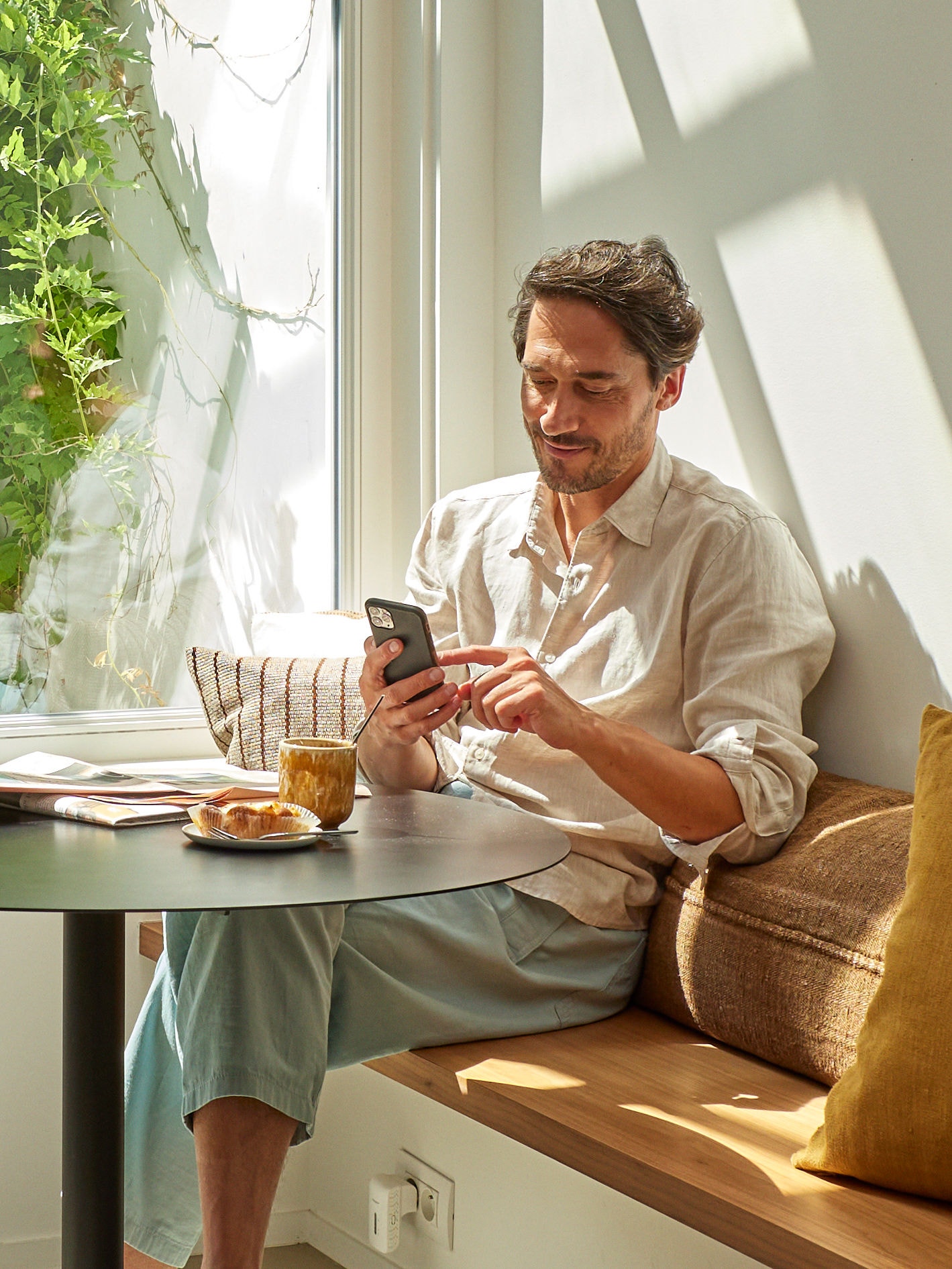 Een ontspannen man die naar zijn telefoon kijkt aan een tafeltje met een kop koffie en iets te eten voor zijn neus