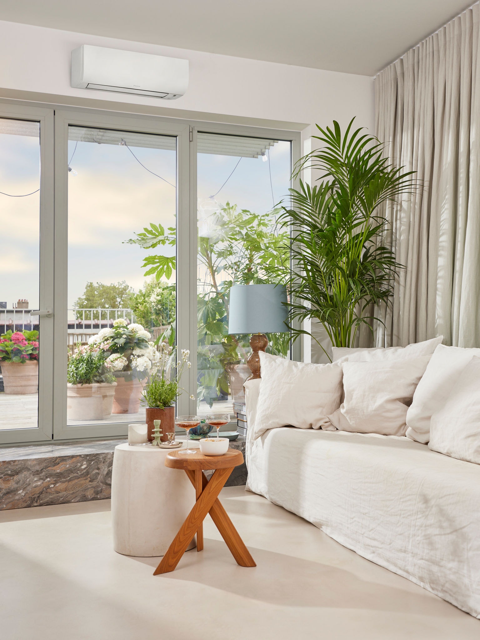 Lichte, gezellige woonkamer met aperitieven op tafel, grote glazen deuren naar een terras met daarboven een airconditioner.