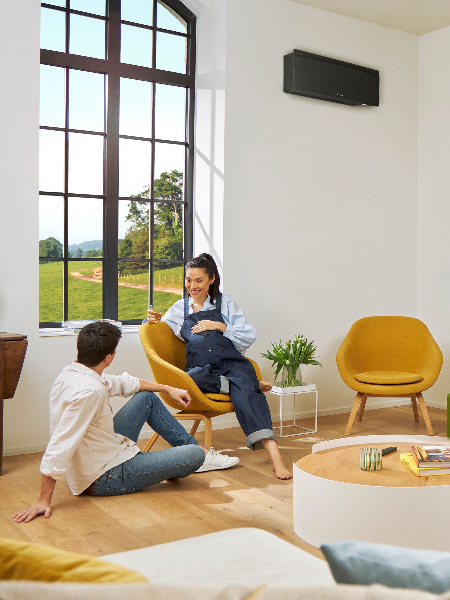 Een man en een vrouw zitten voor een groot raam in een lichte kamer, met een zwarte wandgemonteerde warmtepomp achter hen