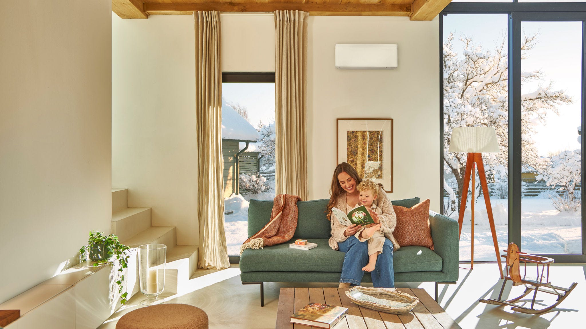 Moeder zit op de bank onder een lucht/lucht warmtepomp aan de wand en leest voor uit een boek voor haar kind dat op haar schoot zit. Ze genieten van de warmte terwijl het buiten sneeuwt.